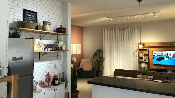 A modern kitchen with white brick walls, a built-in fridge, and shelves with kitchenware. There is a dining area with a sofa and a TV. - House with pool in Caraguatatuba, rustic and modern - Porto Novo beach (Barranco Alto Caraguatatuba)
