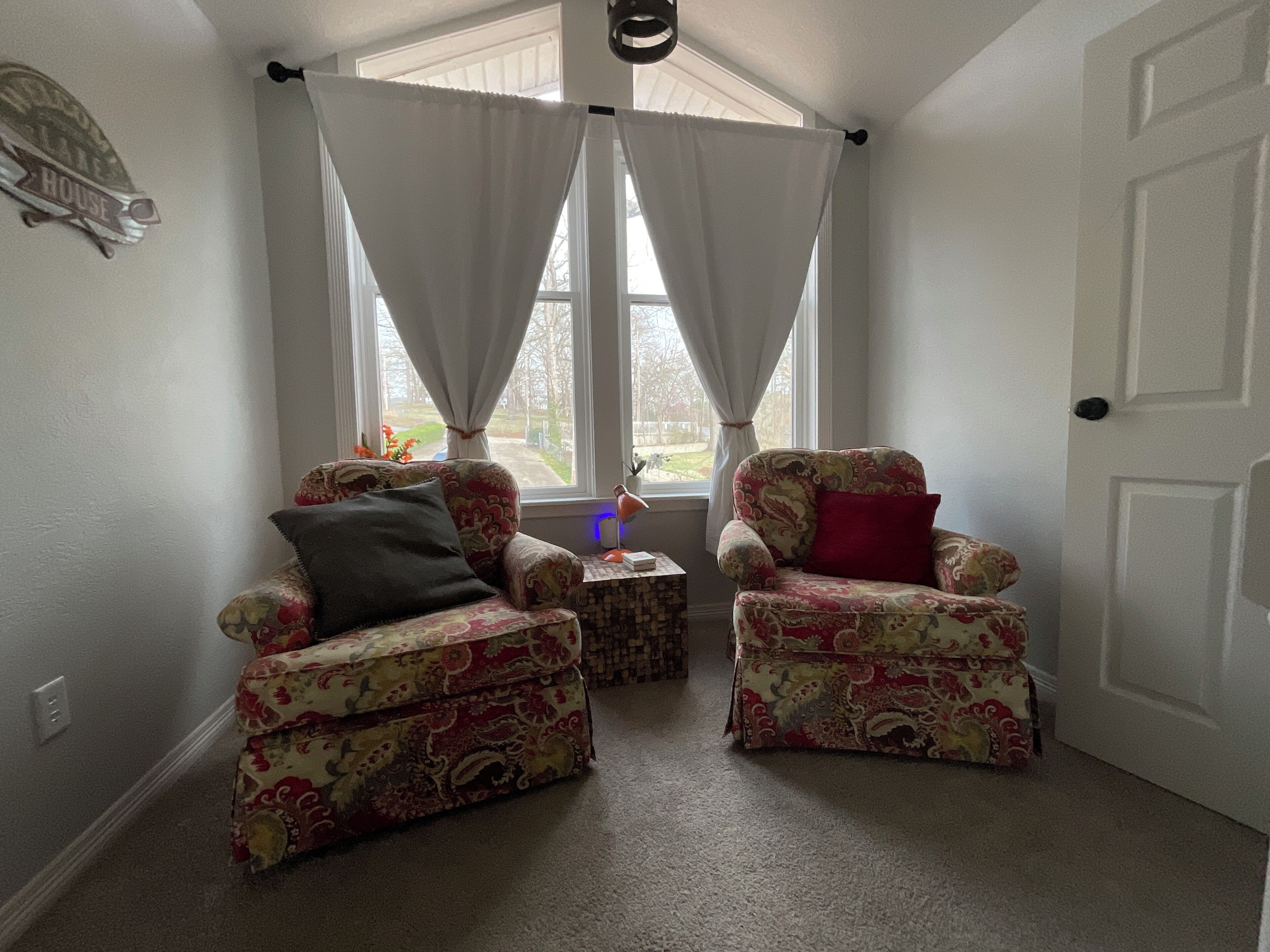Une pièce comprenant deux fauteuils à motifs, une petite table d’appoint et une fenêtre ornée de rideaux blancs.