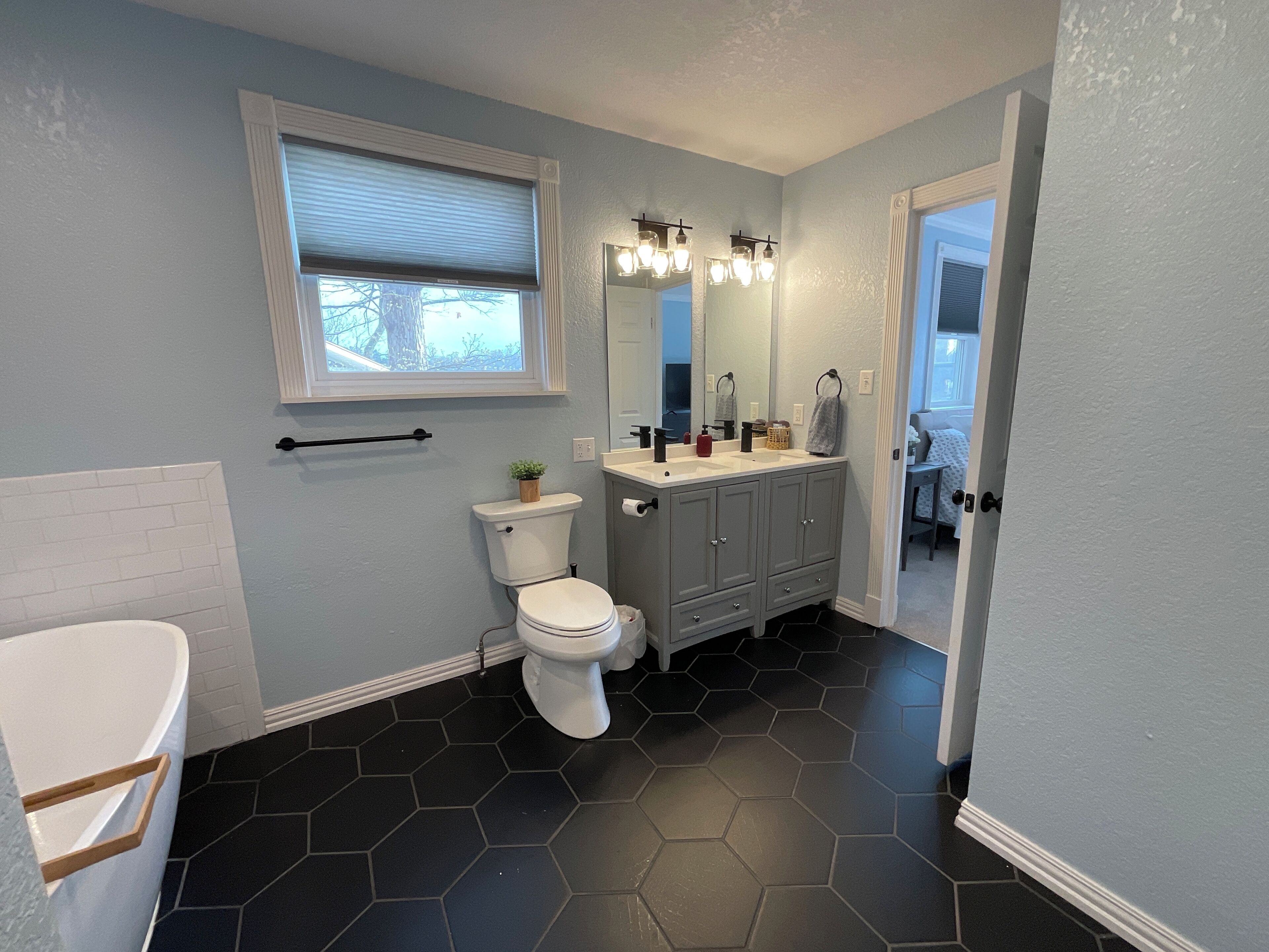 Une salle de bain avec des toilettes, un lavabo et un meuble-lavabo. Il y a une fenêtre avec des stores, un porte-serviettes et une plante posée sur le réservoir des toilettes. Le sol est recouvert de carreaux hexagonaux noirs.