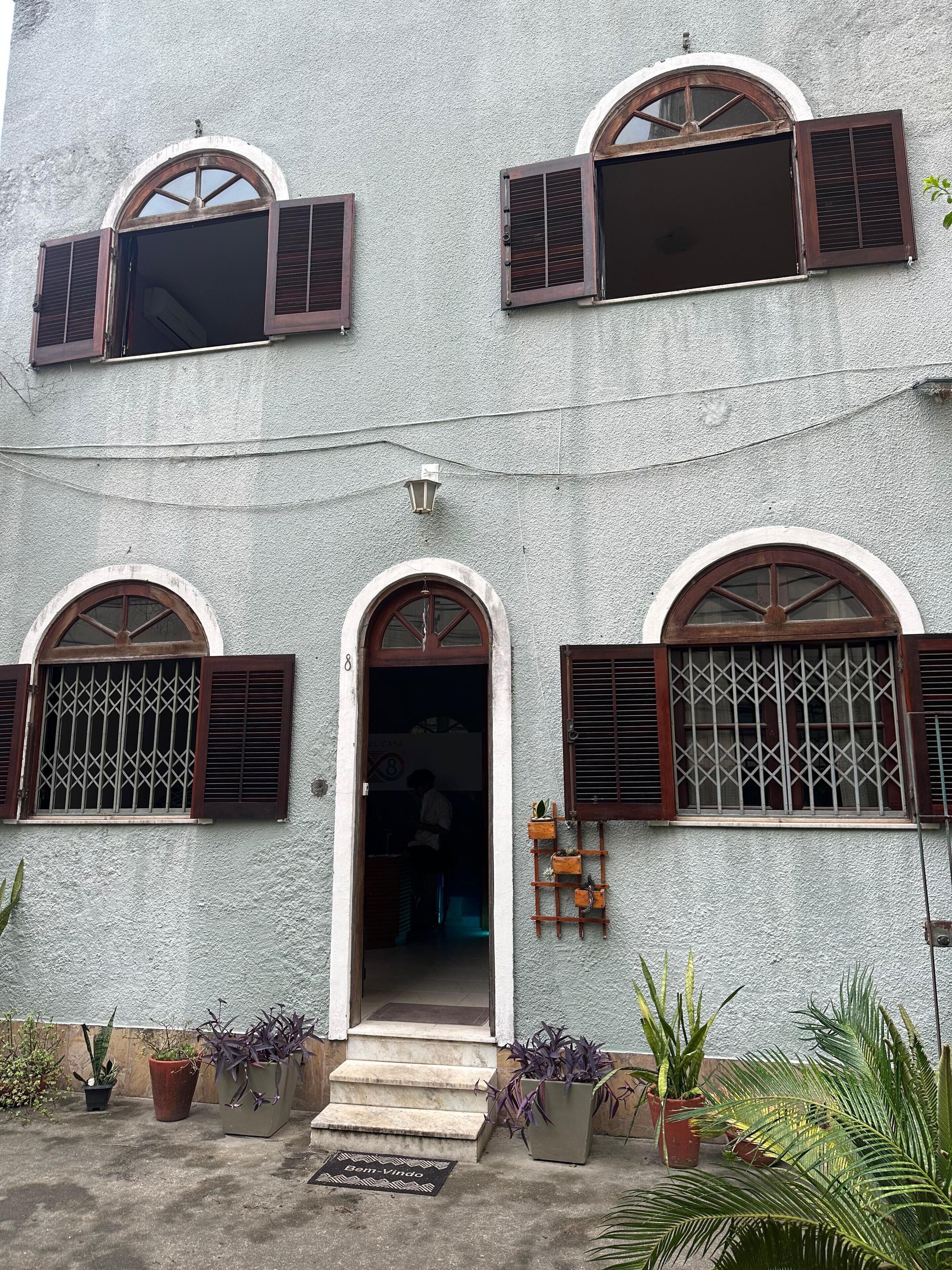 Un edificio de dos pisos con ventanas arqueadas y contraventanas, una pequeña entrada con escalones y plantas en macetas alrededor de la entrada.