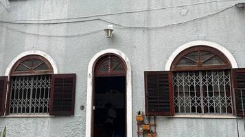 Un edificio de dos pisos con ventanas arqueadas y contraventanas, una pequeña entrada con escalones y plantas en macetas alrededor de la entrada.