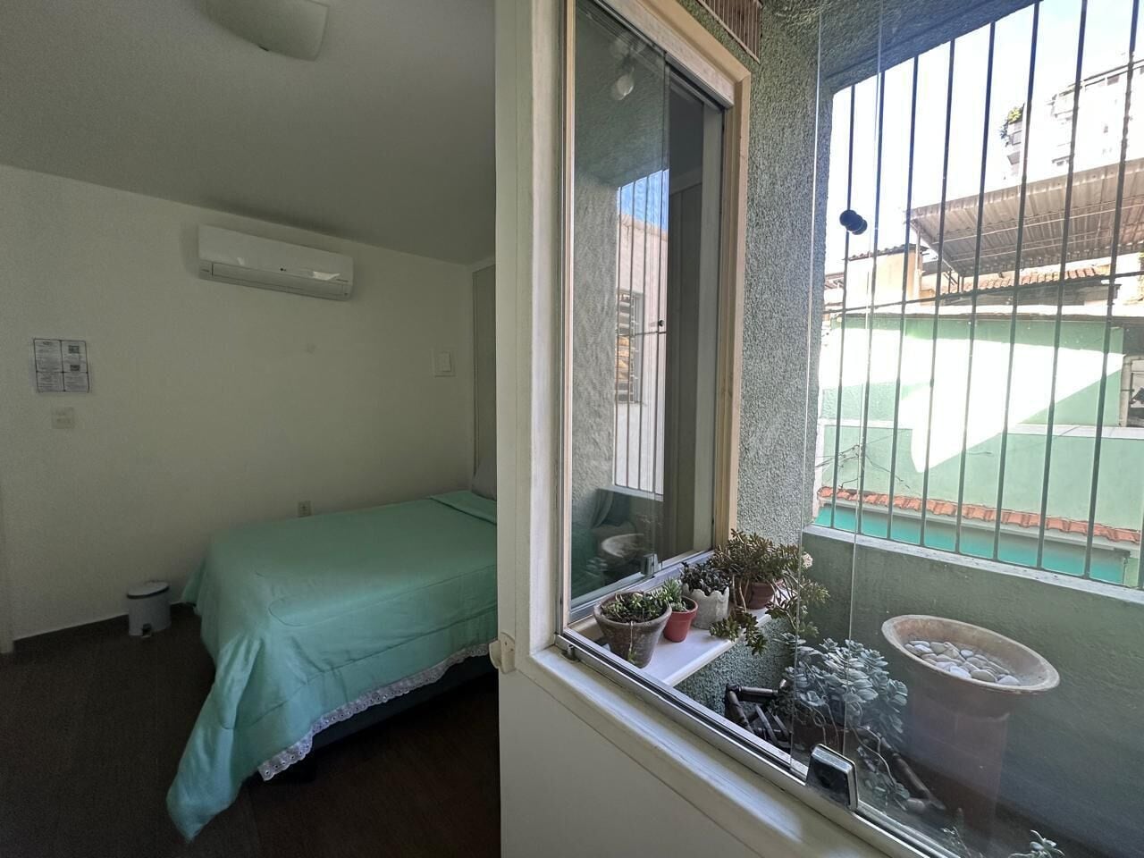 Um quarto pequeno com uma cama de solteiro, uma janela com vista para um prédio e um vaso de planta no parapeito.