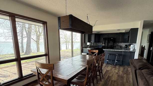 A modern kitchen with dark cabinets, a wooden dining table, and a large window overlooking a body of water.
