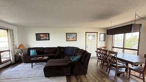 A living room with a sectional sofa, a dining table with chairs, and a large window.