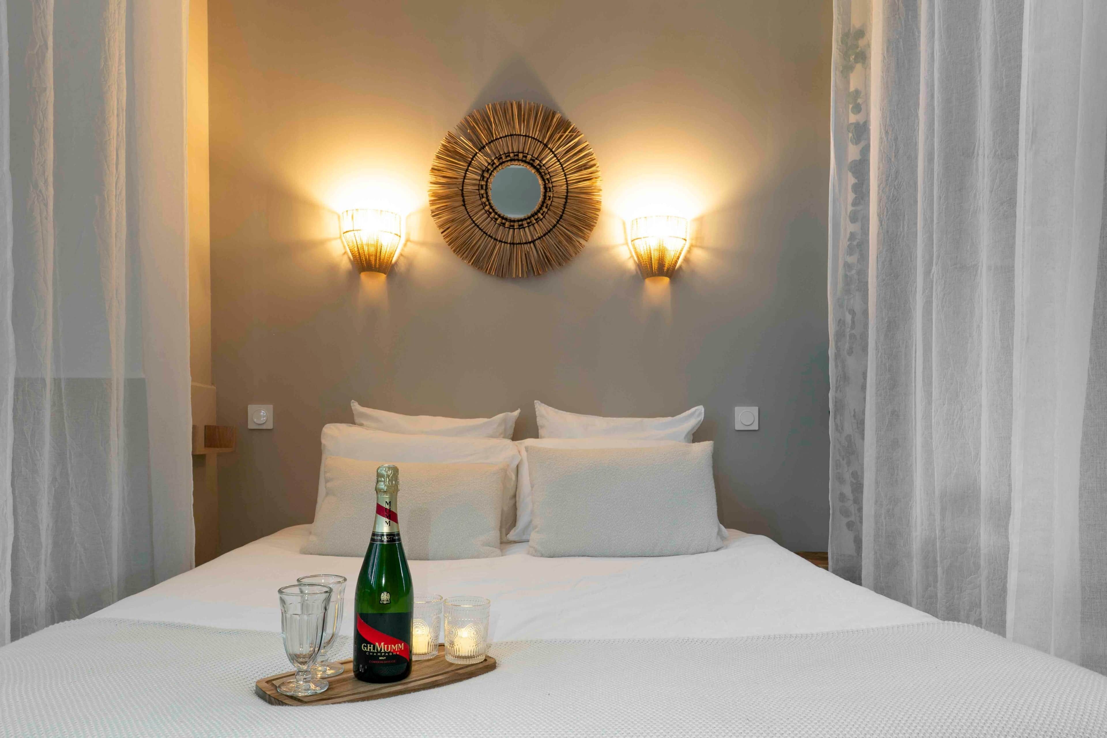 A neatly made bed with white linens, a bottle of champagne, and glasses on a tray, with wall-mounted sconces and a decorative mirror above.