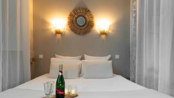 A neatly made bed with white linens, a bottle of champagne, and glasses on a tray, with wall-mounted sconces and a decorative mirror above.