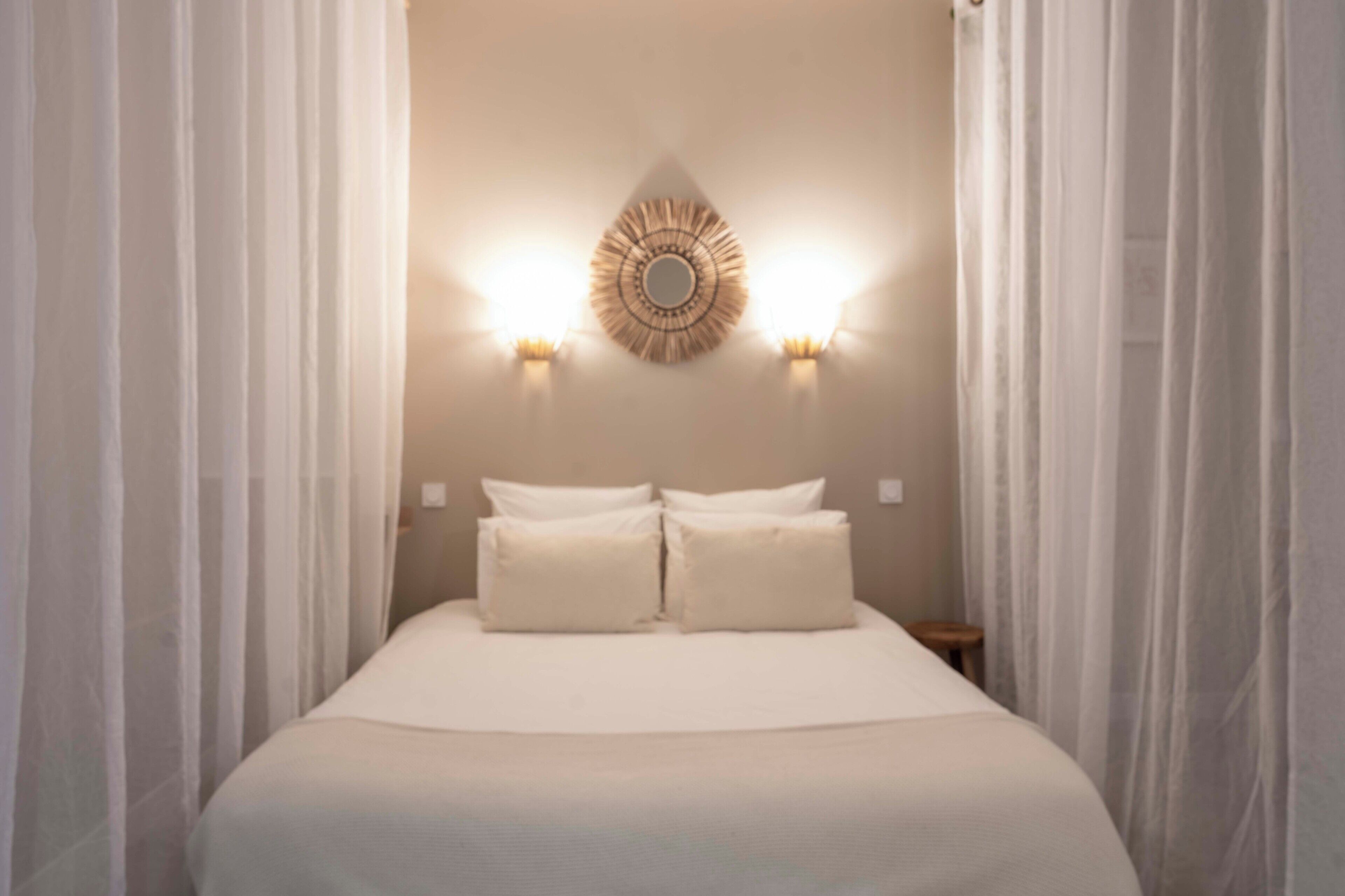 Un letto rifatto con lenzuola bianche e due cuscini, un lampadario a parete e tende di voile bianche.