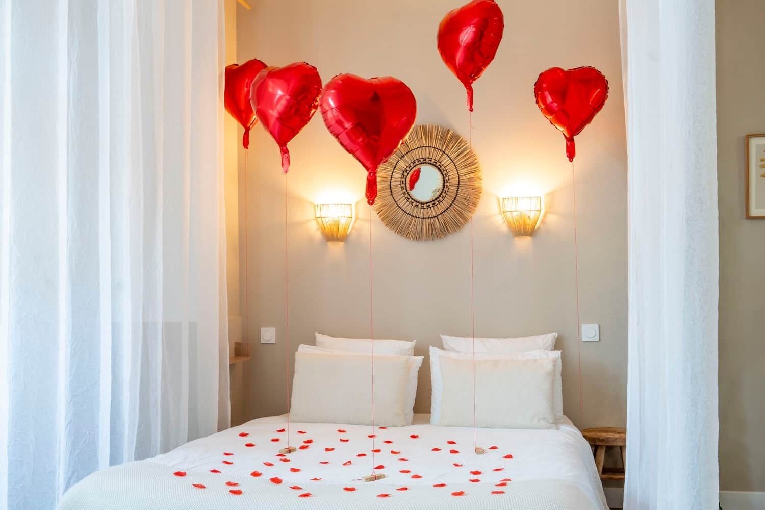 A hotel room with a bed, white curtains, and red heart-shaped balloons.