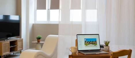 Una sala de estar moderna con un sofá, una mesa de madera con una computadora portátil, un televisor y una ventana con cortinas translúcidas.