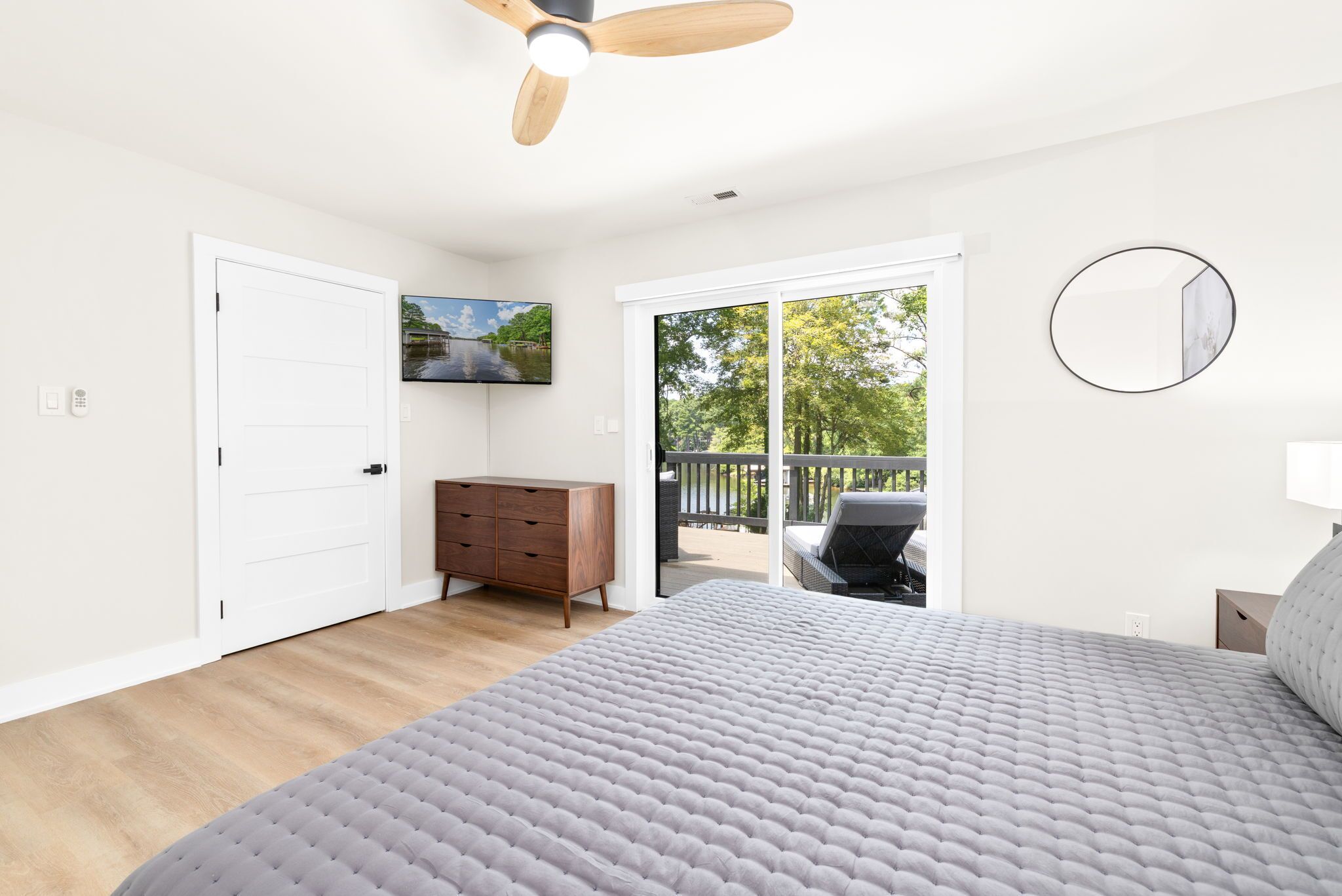A bedroom with a large bed, a ceiling fan, a wooden dresser, a TV, and a balcony with outdoor furniture.