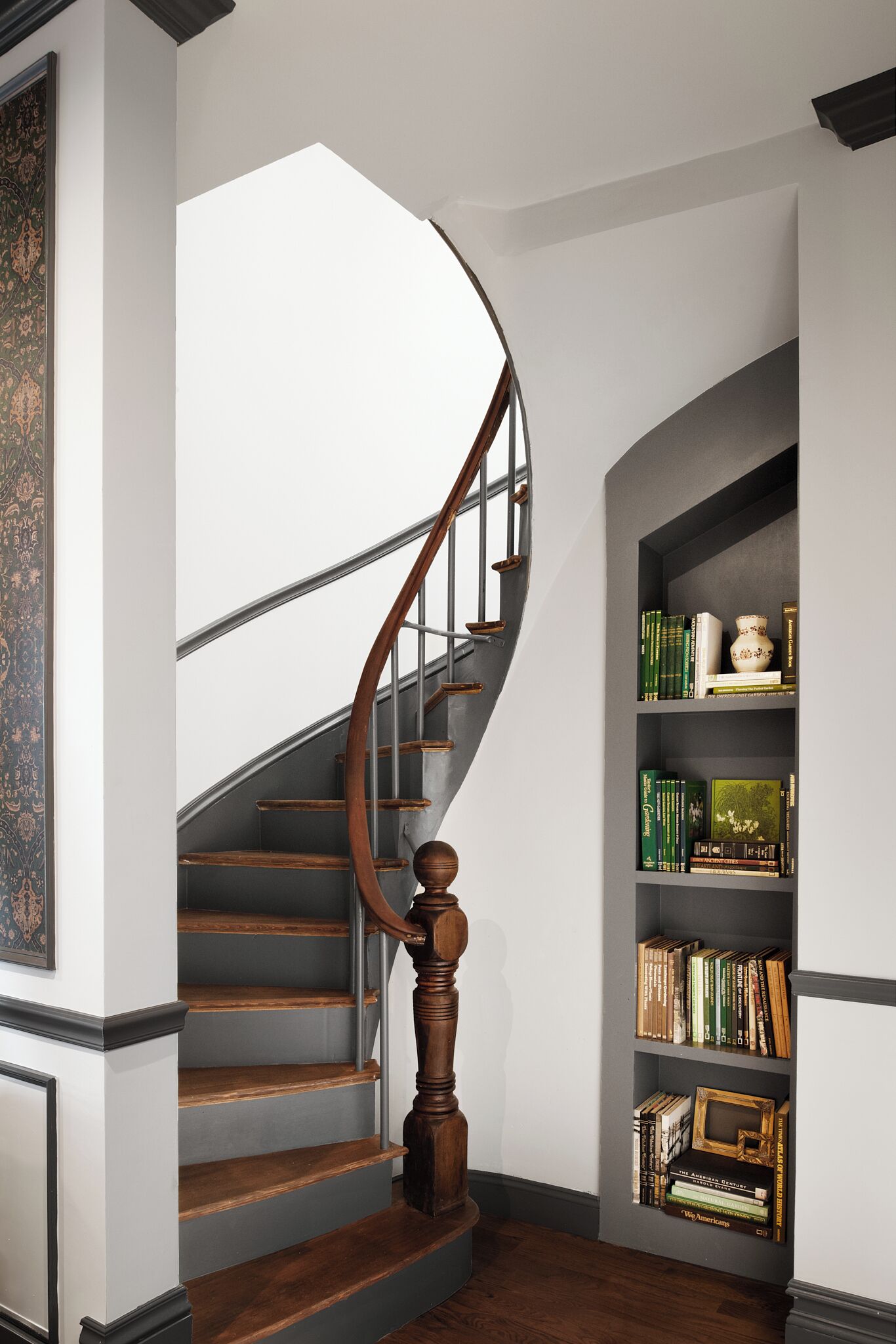 Una escalera con barandilla de madera y suelo oscuro de madera, que conduce a un rellano con una estantería que contiene libros y objetos decorativos.