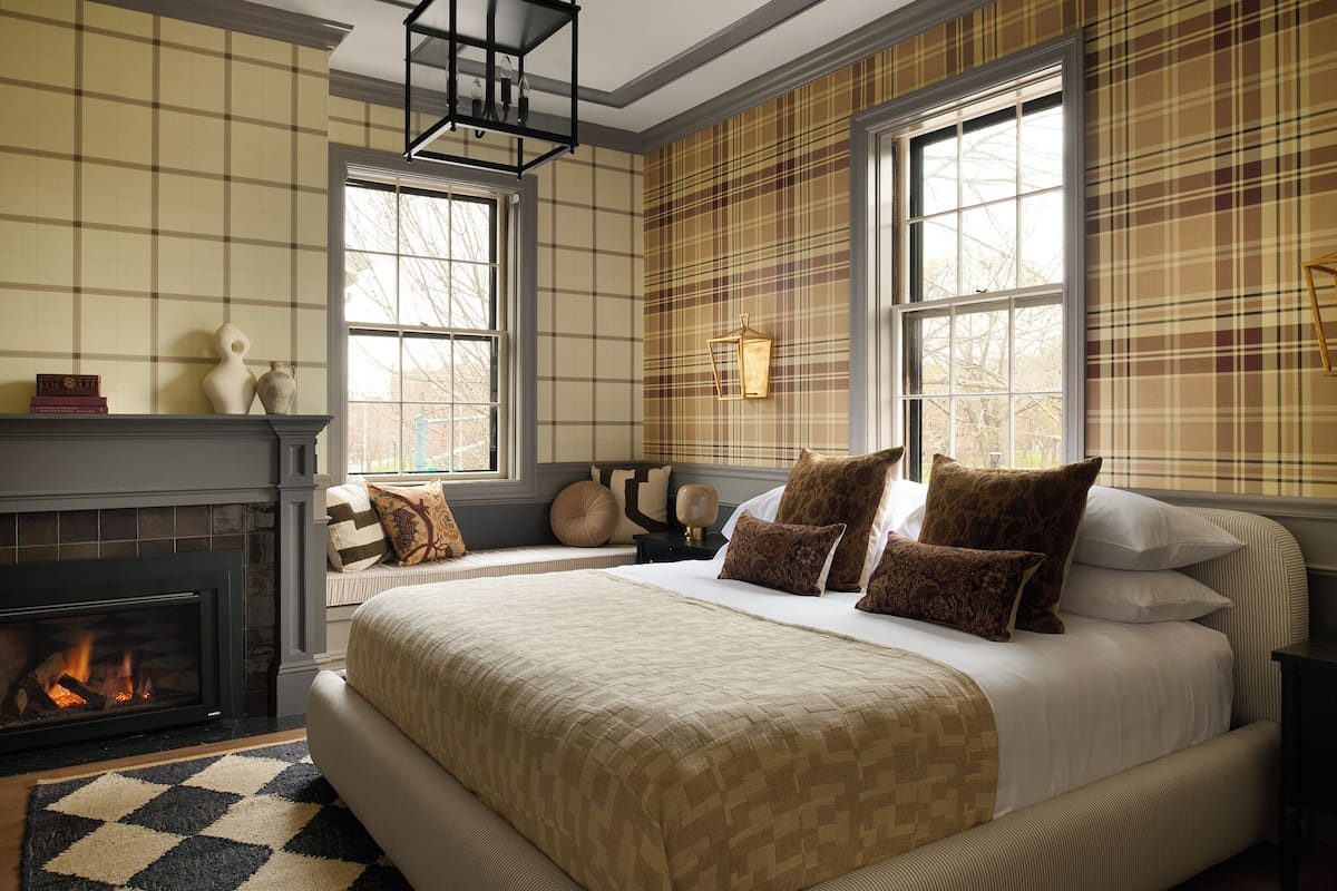 A bedroom with a fireplace, large windows, a bed with multiple pillows, and a patterned rug.