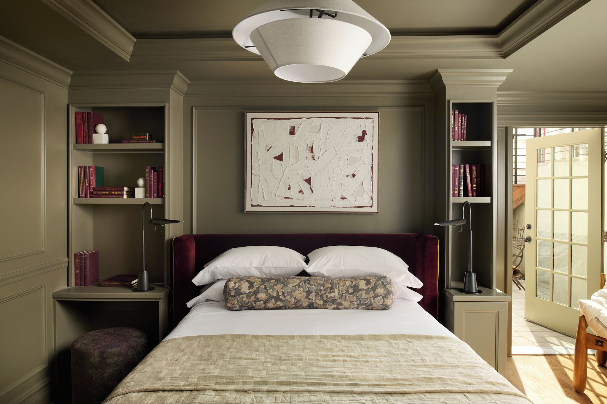A bedroom with a large bed, a bookshelf, a painting, and a door leading to another room.