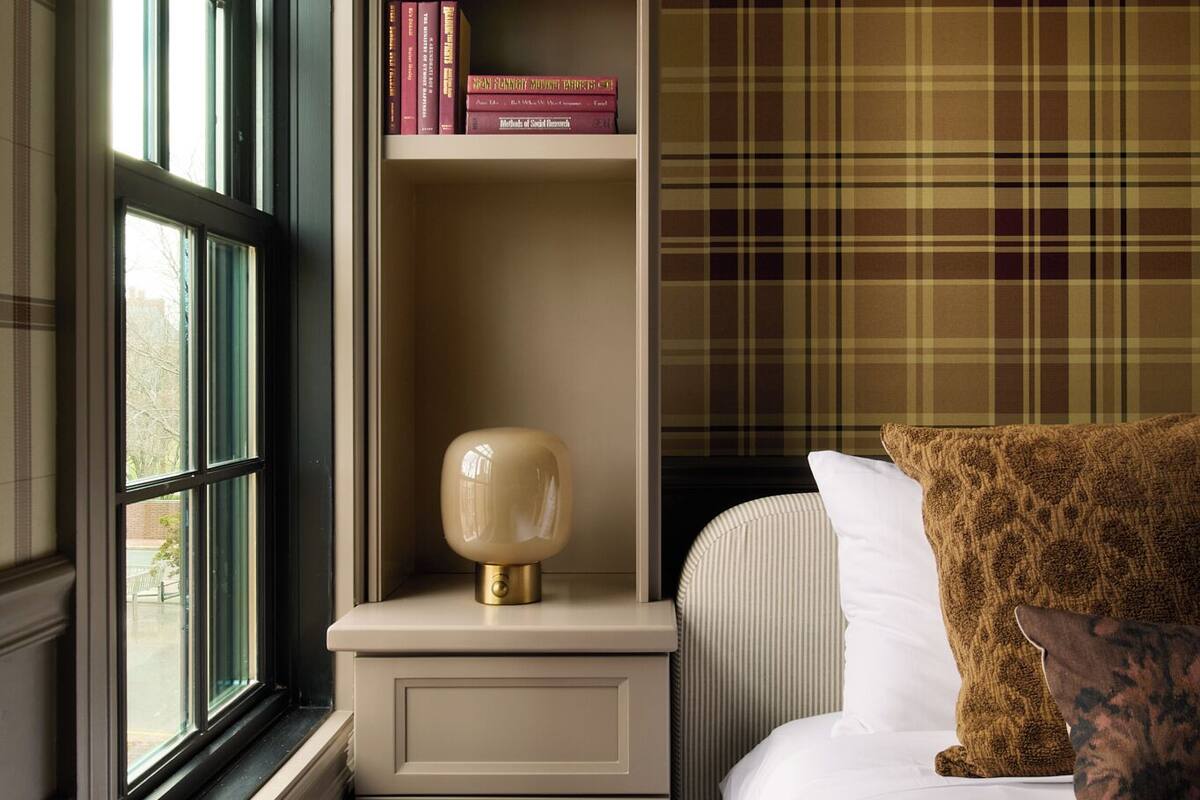 A bedroom with a window, a bedside table with a lamp, a bookshelf, and a plaid-patterned wall.
