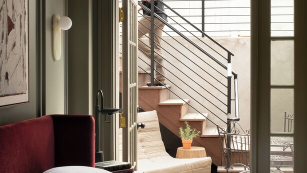 A bedroom with a bed, a red chair, a small table with a book, a potted plant, and a staircase leading to an outdoor area. - Cambridge Common House (Cambridge)