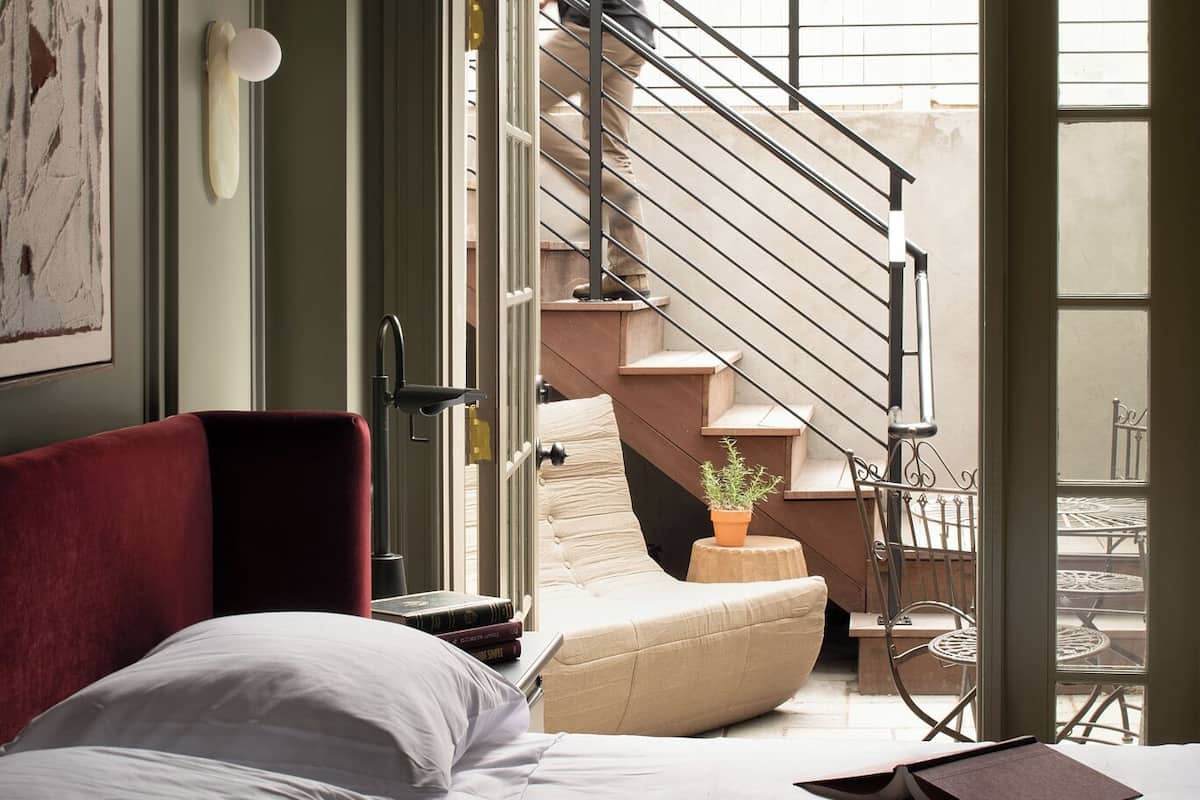 A bedroom with a bed, a red chair, a small table with a book, a potted plant, and a staircase leading to an outdoor area.