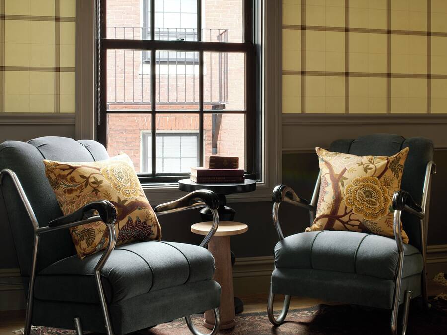 Two armchairs with patterned cushions, a small round table with a book, and a window with a view of a brick building.