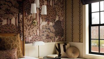 A bedroom with a large bed, a nightstand, a lamp, a window with a view, and a wall with a nature-themed wallpaper.