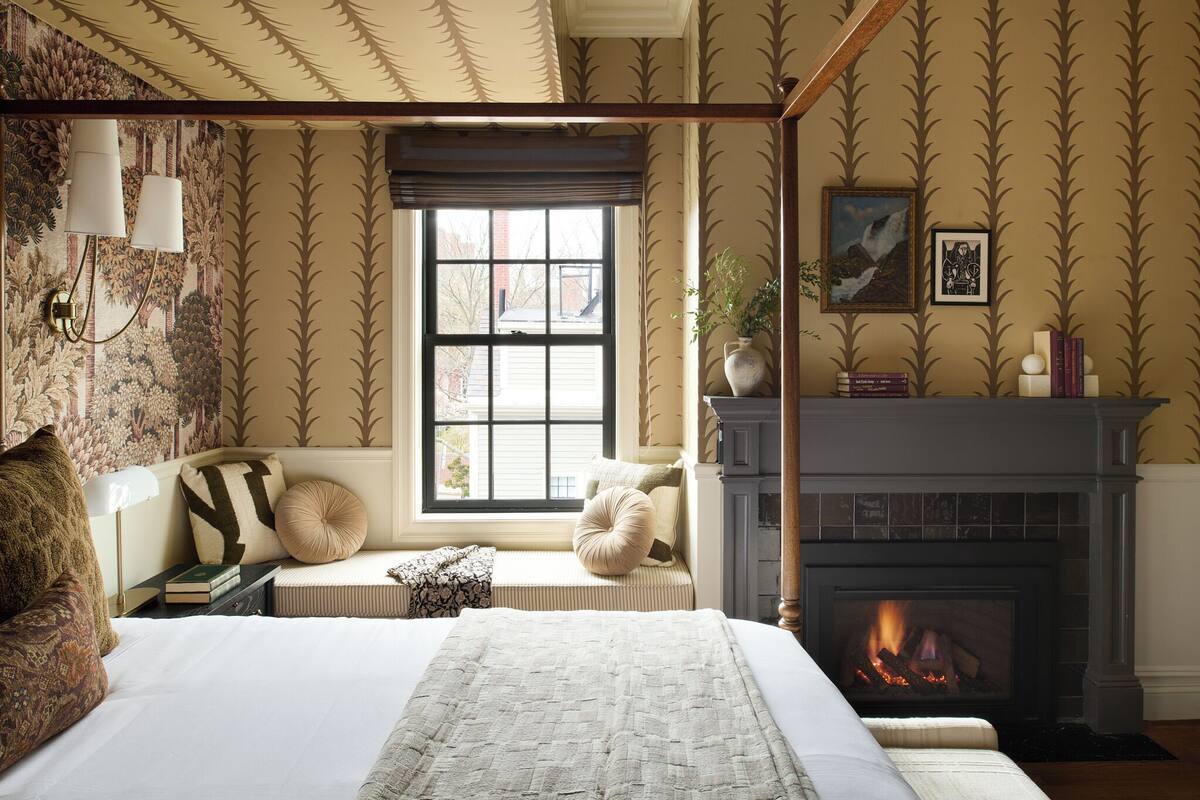 A bedroom with a four-poster bed, a fireplace, a window with blinds, and a bookshelf.