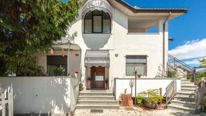 A two-story house with a white exterior, a large arched window, and a front door. There are steps leading to the entrance, and a small garden area with potted plants.
