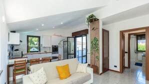 A modern living room with a sofa, a dining area with chairs, a kitchen with cabinets and appliances, and a large window.