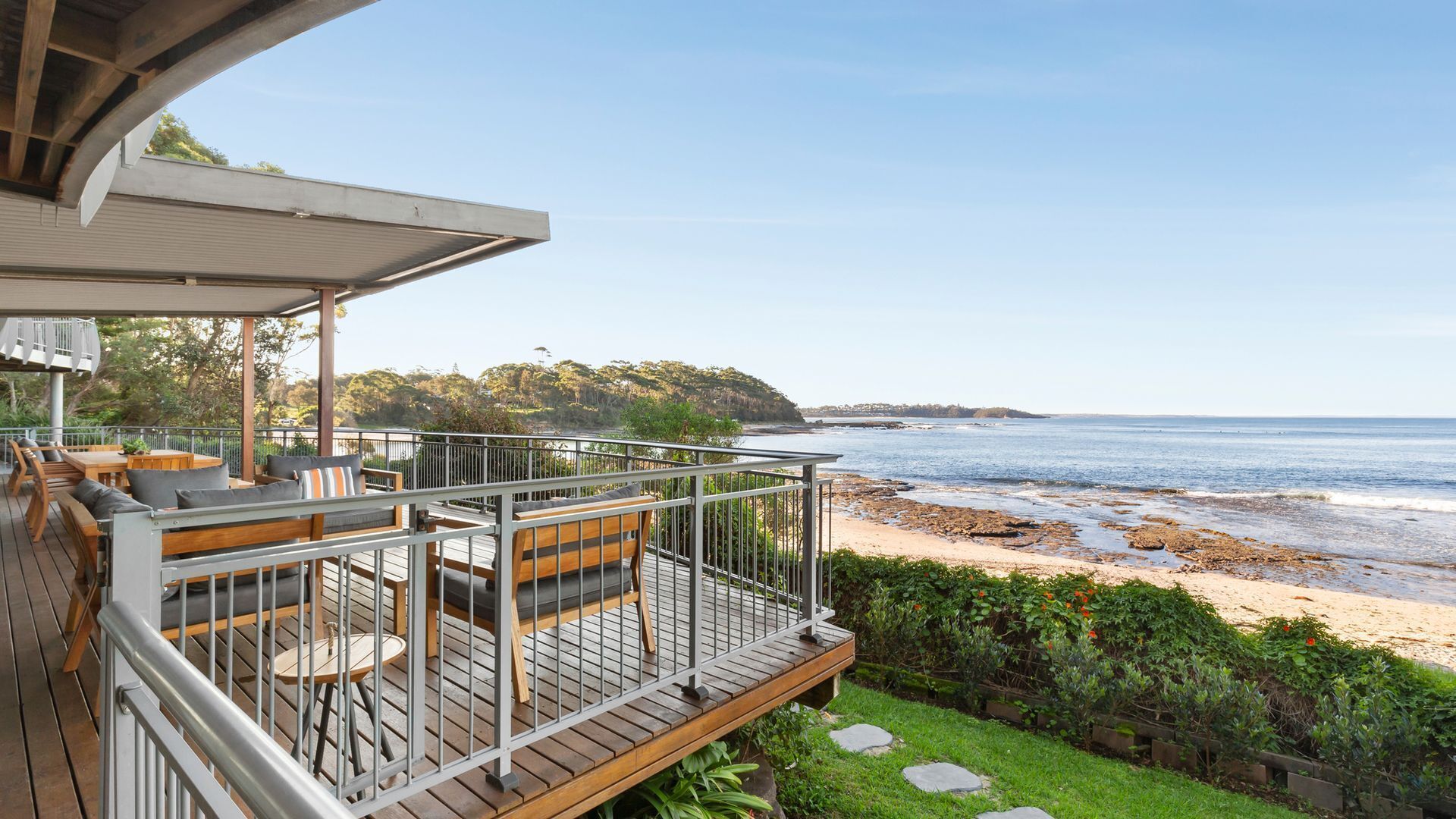 A wooden deck with outdoor furniture, overlooking a beach with rocky shoreline and ocean views.