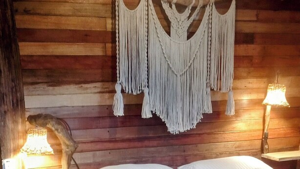 A cozy wooden cabin room with a bed, macramé wall hanging, and two bedside lamps. - Cabin in the Salento cloud forest (Salento)