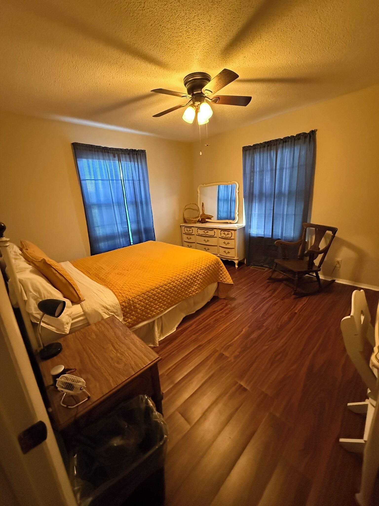 A bedroom with a bed, a dresser, a chair, a ceiling fan, and a window with curtains.