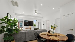 A modern living room with a grey sofa, a wooden dining table with four chairs, a large potted plant, and a flat-screen TV mounted on the wall.