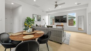 A modern living room with a grey sofa, a wooden dining table with four chairs, a ceiling fan, and a flat-screen TV mounted on the wall.