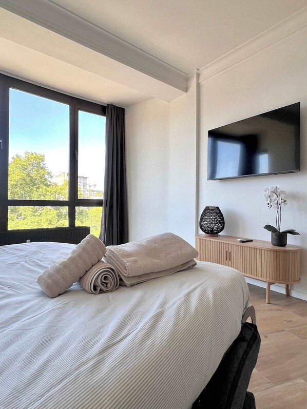 Une chambre moderne avec un grand lit, un téléviseur à écran plat fixé au mur et une vue sur l’extérieur par une fenêtre.