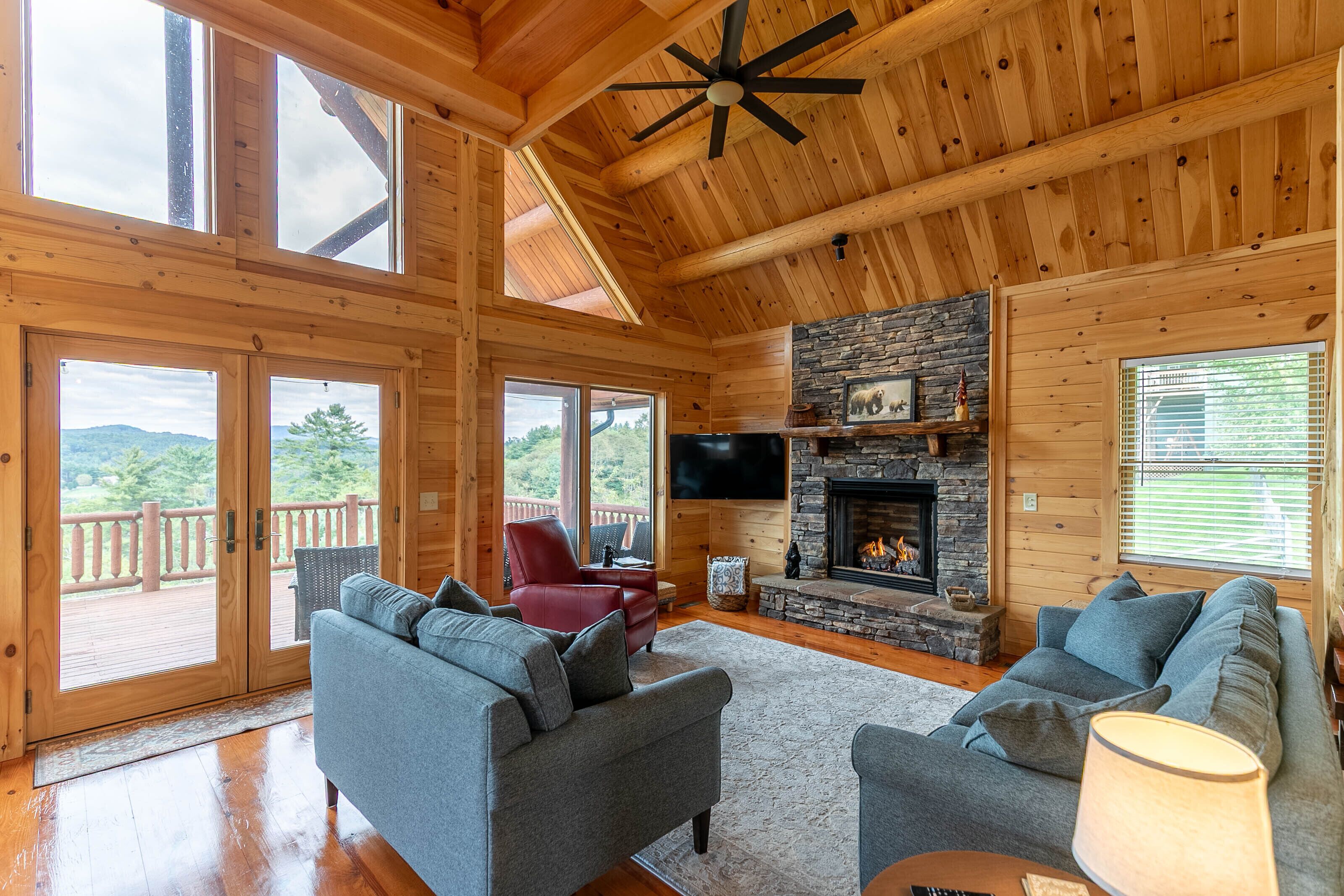 Une cabane en bois spacieuse, dotée d’une cheminée en pierre, de deux canapés, d’un fauteuil rouge et d’un grand téléviseur.