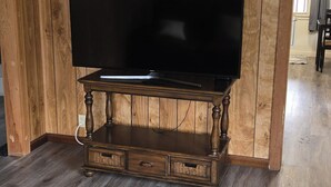 Una habitación con paneles de madera, un televisor de pantalla plana grande sobre un soporte de madera, una alfombra estampada y un vano que conduce a otra habitación.