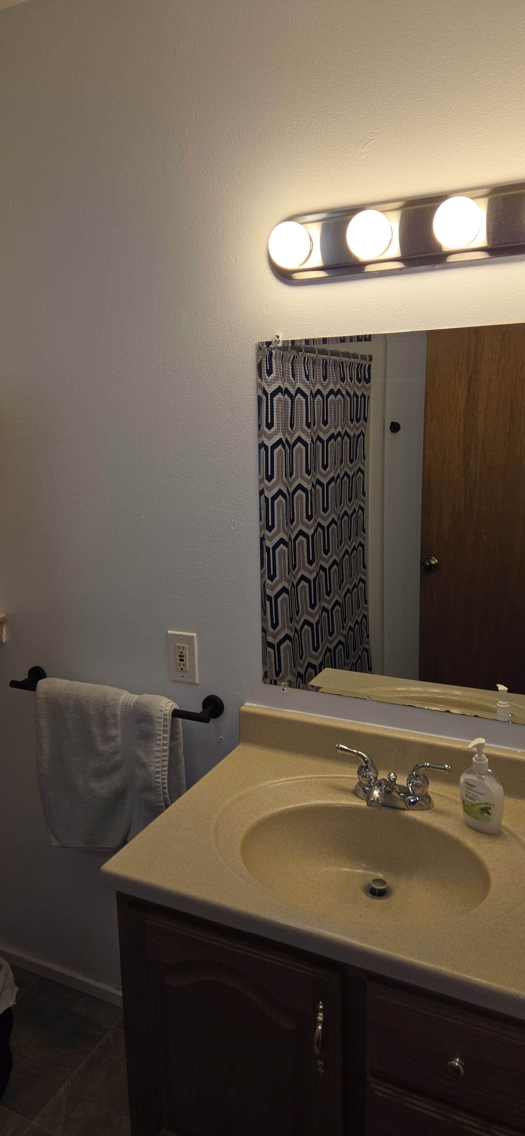 A bathroom with a beige sink, a mirror with lights, a towel rack, and a patterned curtain.