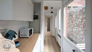 Un estrecho pasillo con suelo de madera, paredes blancas y una ventana con vista a un muro de ladrillo. Se ve un microondas y un electrodoméstico azul a la izquierda.