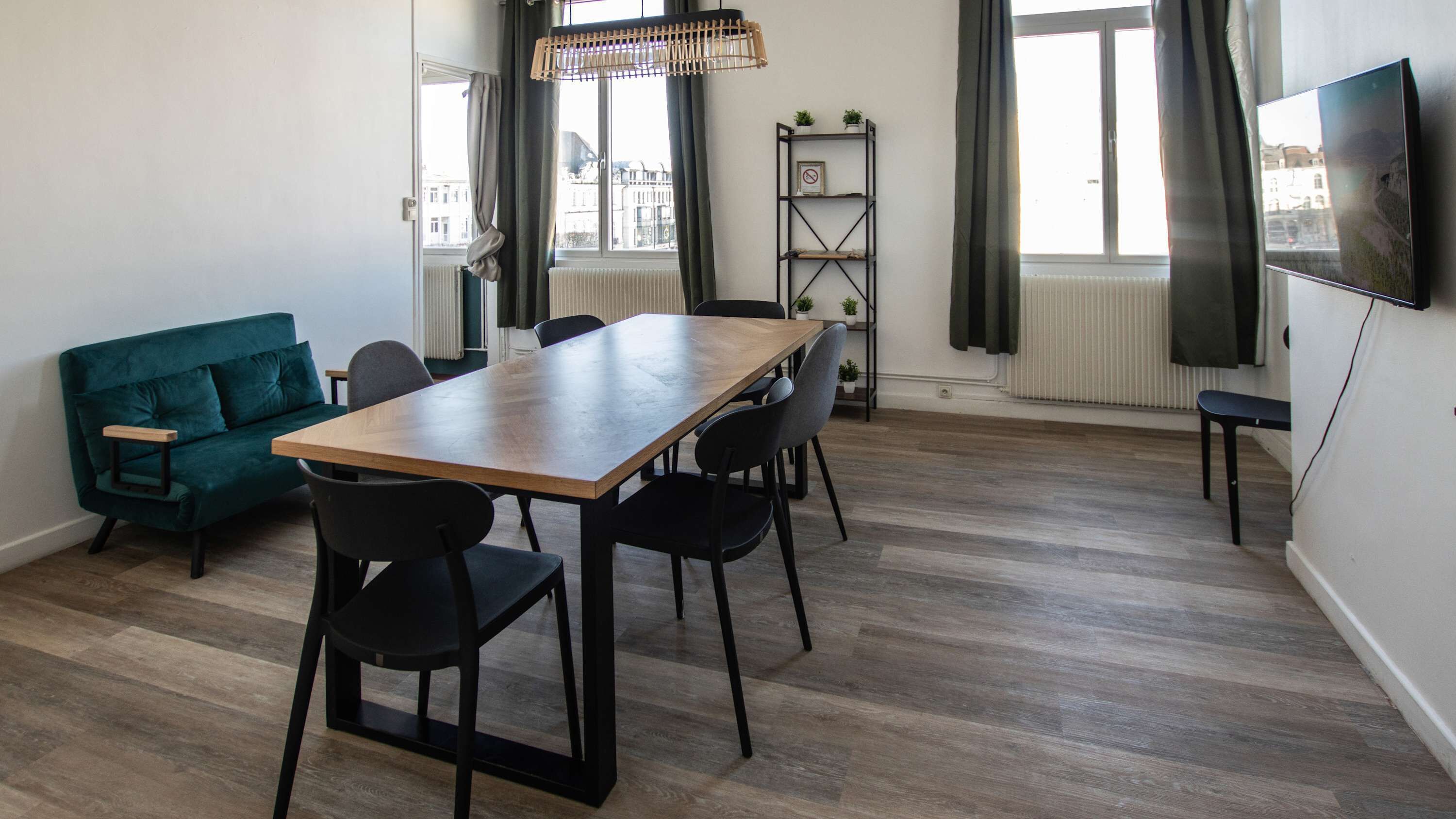 A modern living room with a wooden dining table, black chairs, a teal sofa, and a TV mounted on the wall.