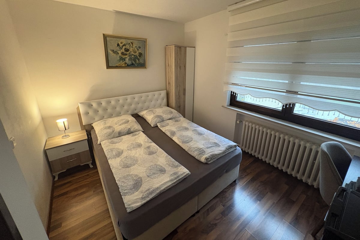 A bedroom with a bed, a nightstand, a radiator, a window with blinds, and a painting on the wall.