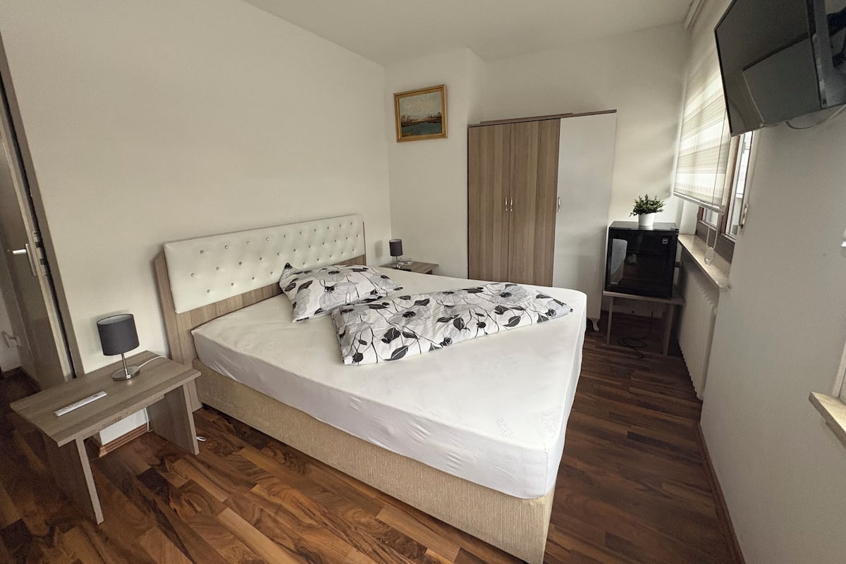 A bedroom with a bed, a nightstand, a wooden wardrobe, a small table with a plant, and a TV mounted on the wall.