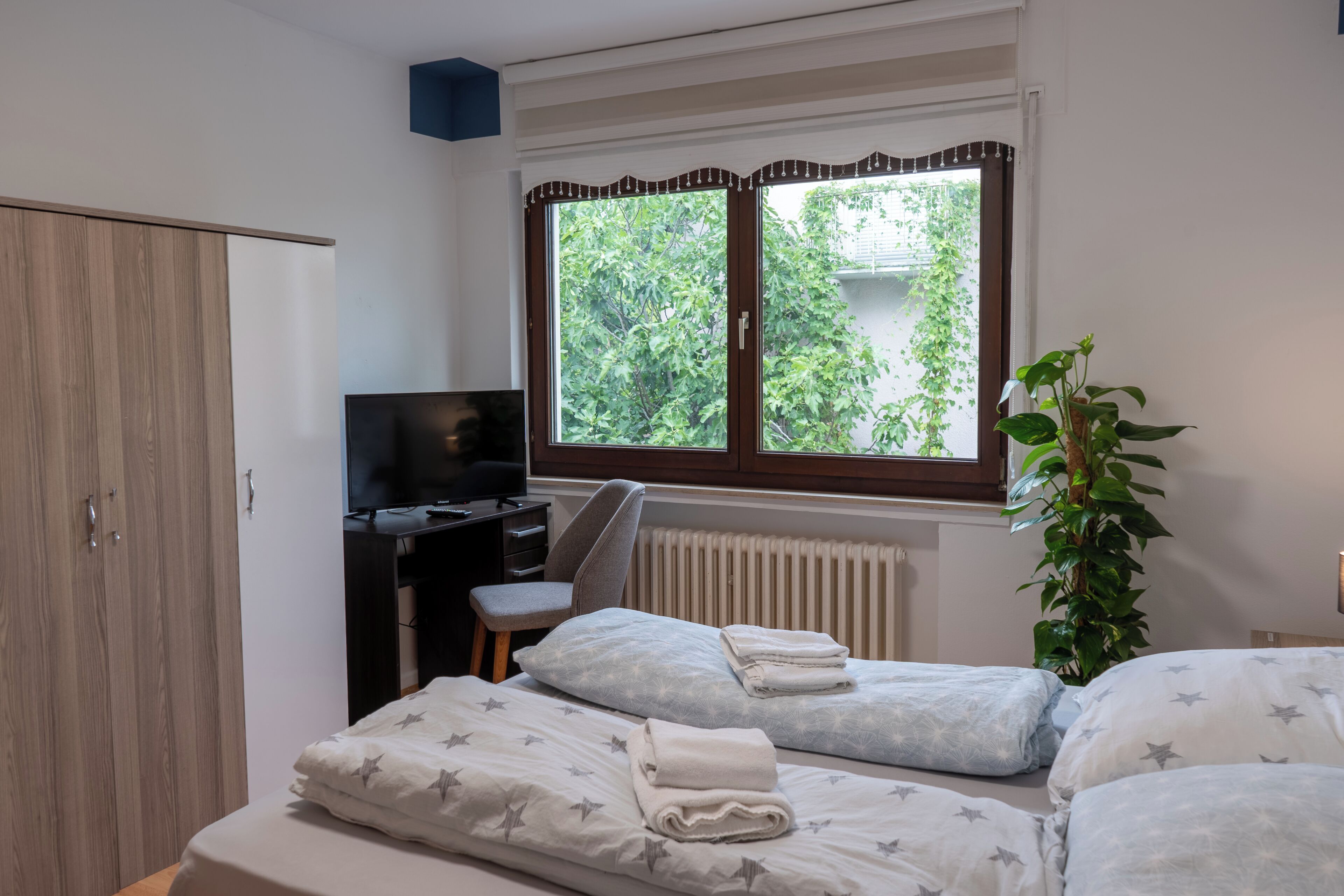 Ein Schlafzimmer mit zwei Betten, einem Schreibtisch, einem Fernseher und einem Fenster mit Blick auf Grünflächen.