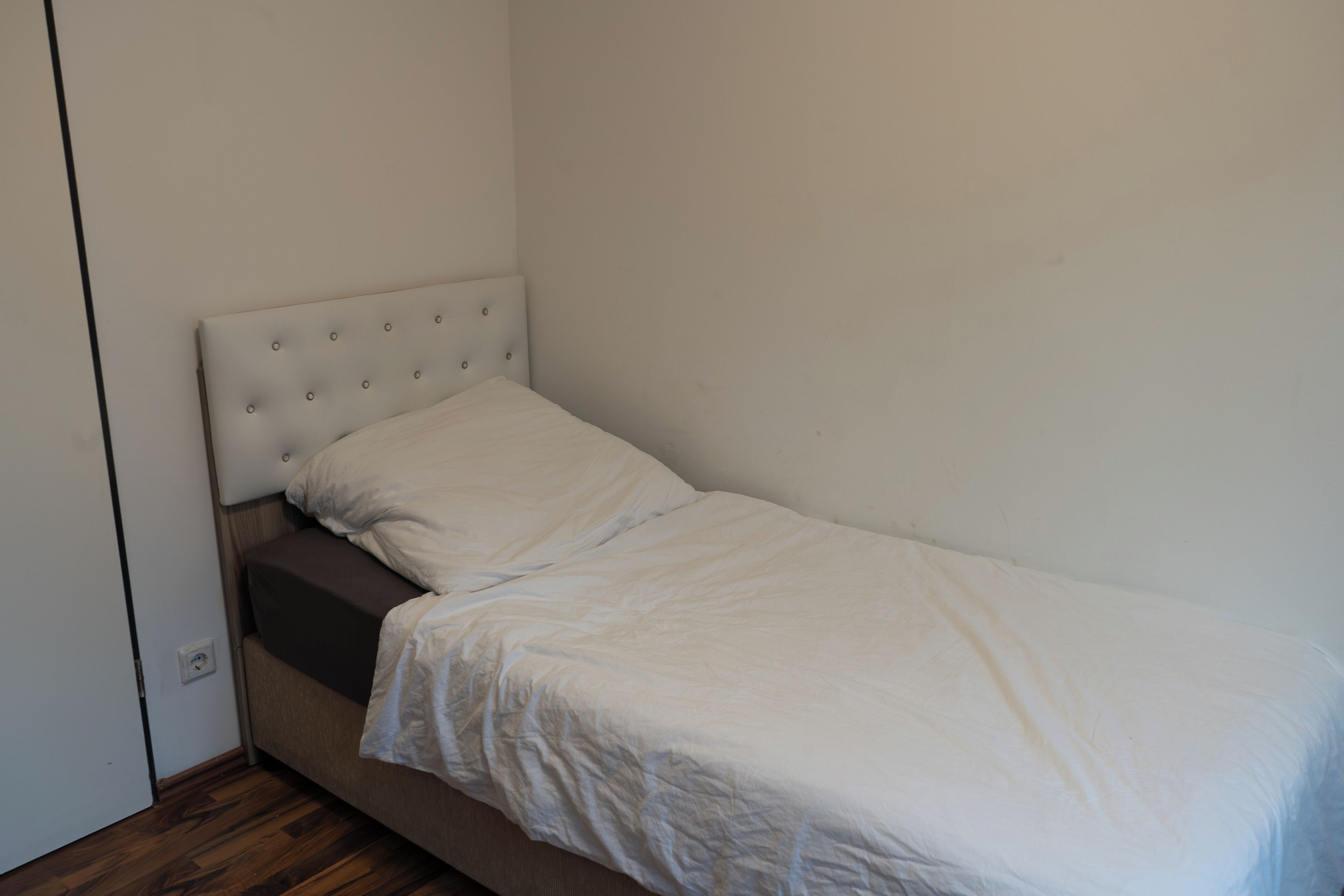 A single bed with a headboard and a mattress in a room with wooden flooring and white walls.