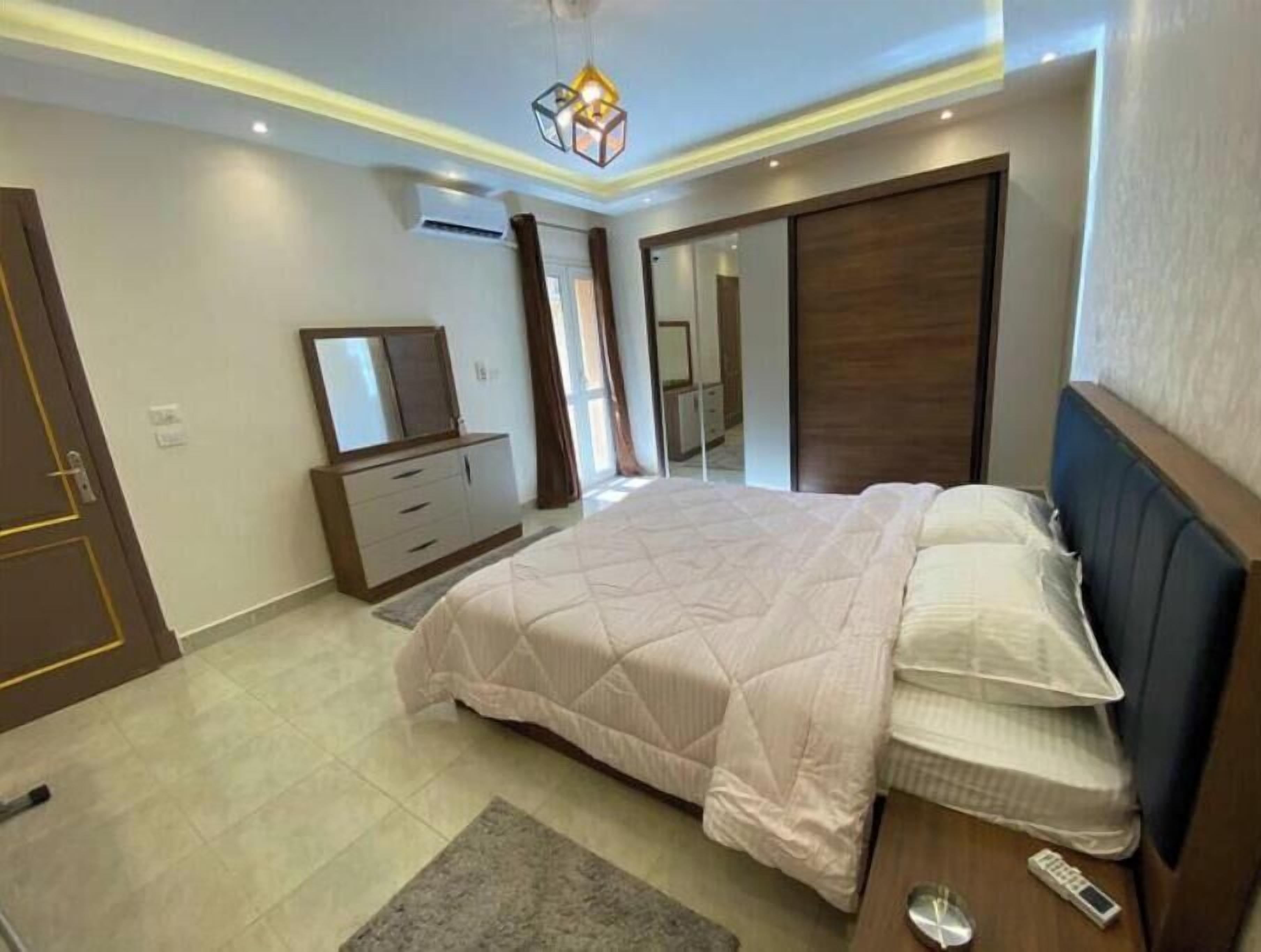 A modern bedroom with a large bed, a dresser, a mirror, and a wardrobe.