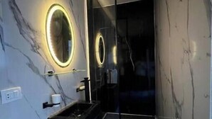 A modern bathroom with a marble wall, a black vanity, a glass shower enclosure, and a circular mirror with a light. - Alrehab cozy apartment (Cairo Governorate)