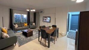 A modern living room with a sofa, coffee table, dining table, and chairs. There is a TV mounted on the wall and a window with curtains. - Alrehab cozy apartment (Cairo Governorate)