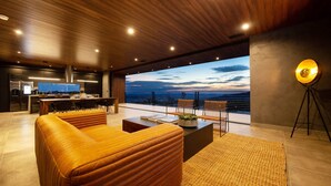 Un salon moderne avec un plafond en bois, une grande fenêtre donnant sur un paysage magnifique, un coin repas et une cuisine équipée d’un téléviseur à écran plat.