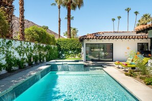 A swimming pool with a tiled edge, a small outdoor seating area with cushions, and a building with a tiled roof and large windows.