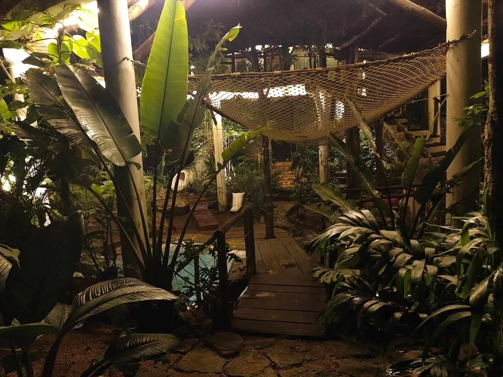 Un sentier en bois mène à un espace de repos abrité, doté d’un toit en fibres tressées, et est entouré d’une végétation tropicale luxuriante.