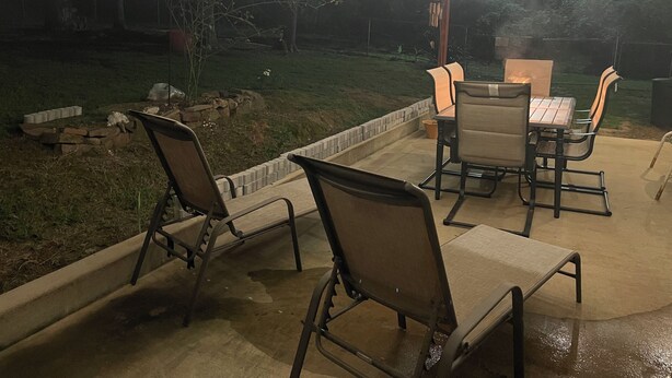 Une terrasse extérieure dotée d’un sol en béton, entourée d’un petit muret en briques. On y trouve quatre chaises en métal et une table, avec une source de lumière placée en dessous.