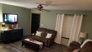 Un salon comprenant une télévision, un ventilateur de plafond, un canapé, une table basse et une fenêtre avec des rideaux.
