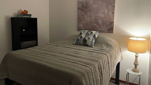 Un lit bien fait, avec une couvre-lit à rayures et un coussin aux motifs géométriques, un tableau abstrait accroché au mur, une petite bibliothèque décorée d’une citrouille et une lampe de table posée sur une table d’appoint.