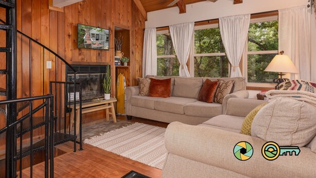 A cozy living room with a wooden interior, a fireplace, a sofa, and a staircase. - Treehouse Mountain Stay - unique cozy quiet! (Basye)